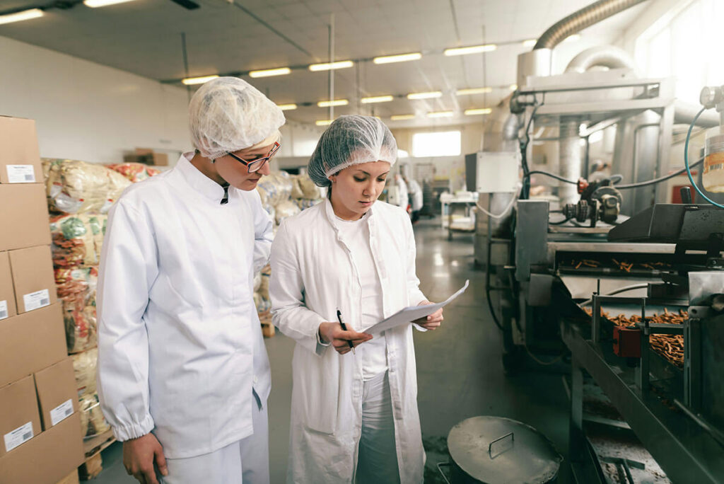 Two women looking at papers standing next to an assembly line of food.